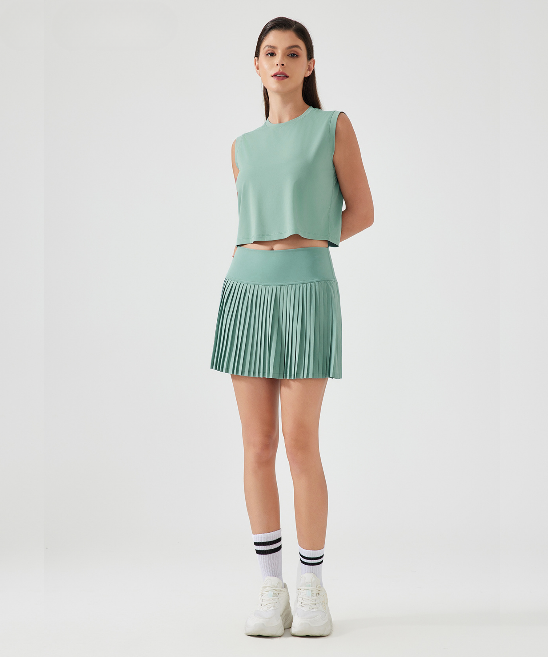 Woman wearing a green sleeveless top and high-rise pleated pickleball skirt on a white background