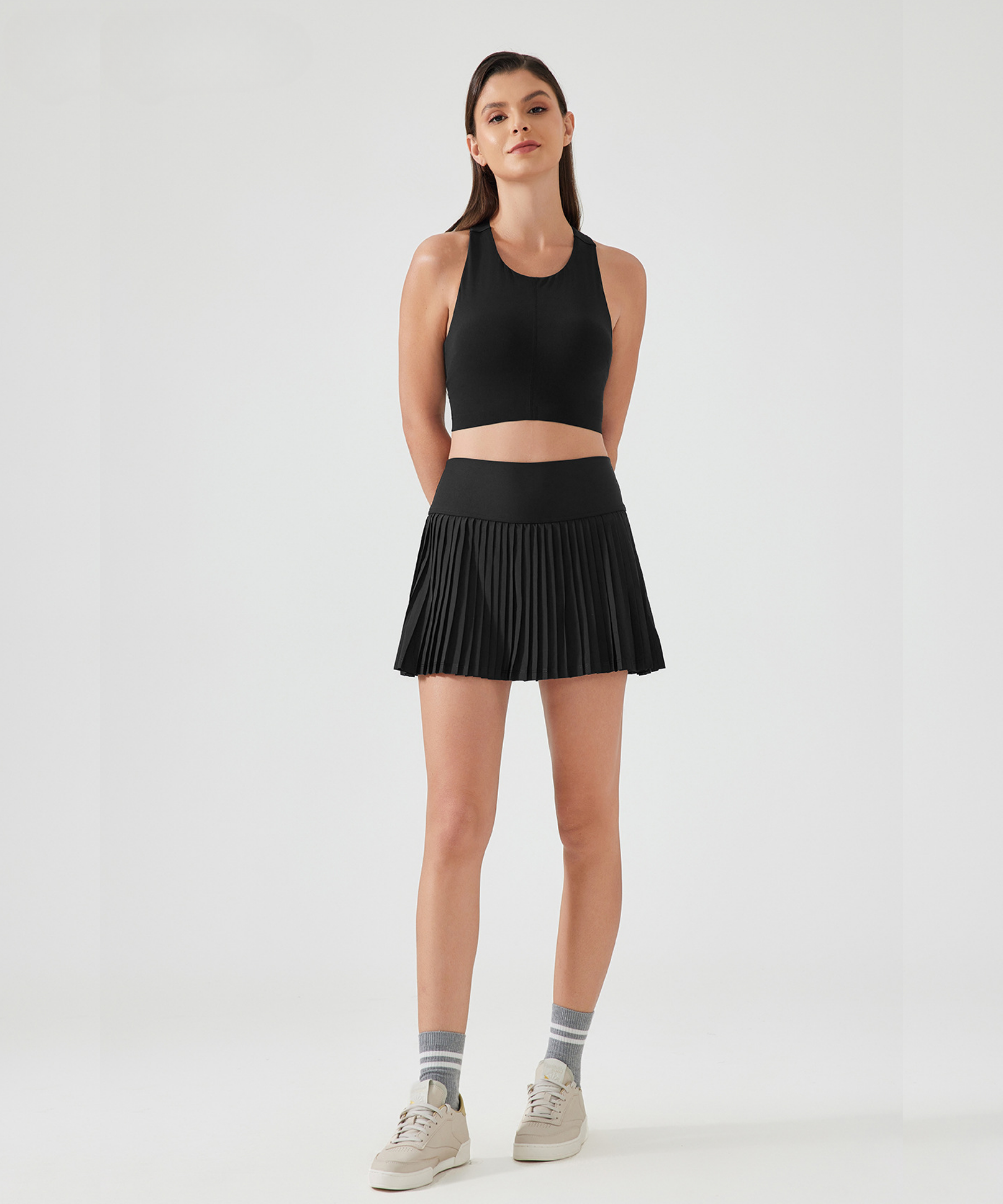 Woman wearing a black athletic tank top and a black high-rise pleated pickleball skirt on a white background
